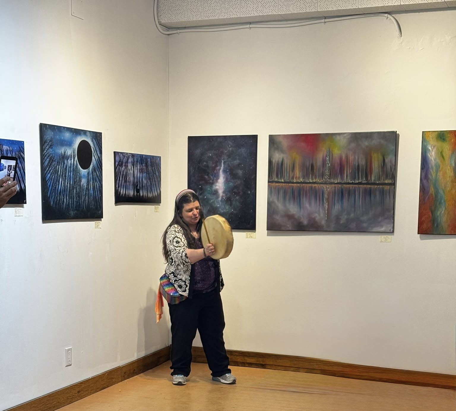 Caroline Little Bear holding a drum in a gallery displaying her paintings